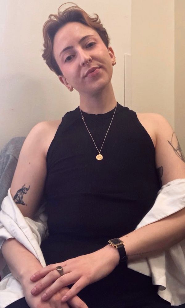 A photo of me, with short red/ginger hair, wearing a black tank top and gold jewelry, sitting with my hands in my lap, looking into the camera  