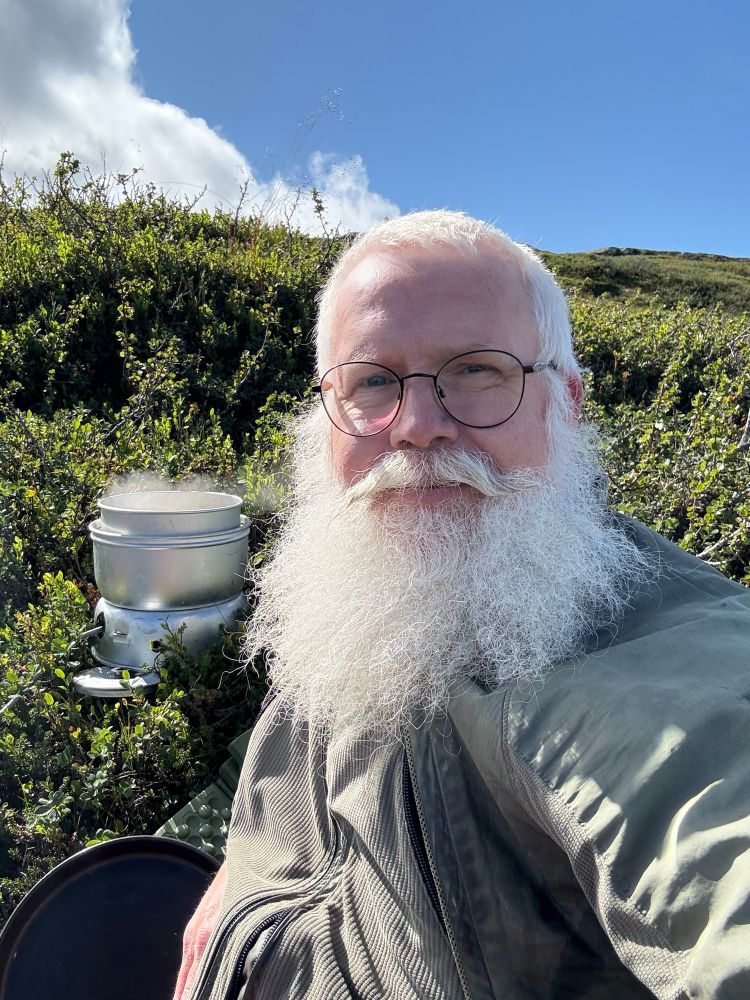 White haired and bearded man sitting by his Trangia cooker
