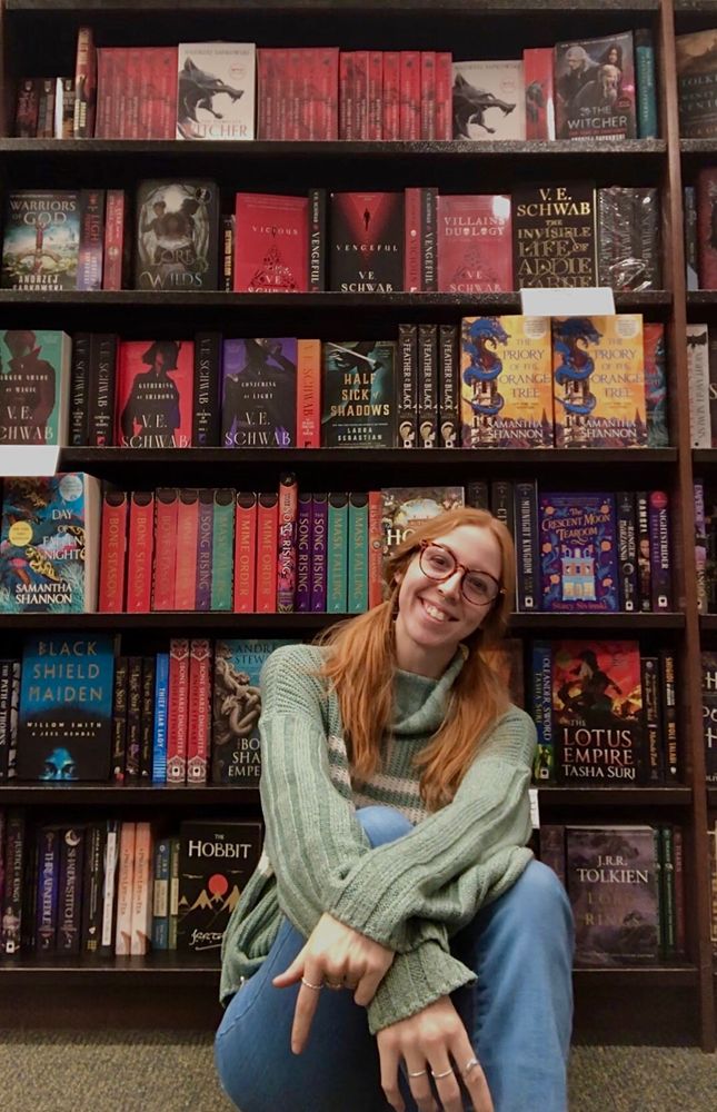 an orange haired woman with glasses sitting in front of large bookshelves, smiling wearing a green sweater and jeans. the books behind her are fantasy titles of all colors, but red sticks out the most. 