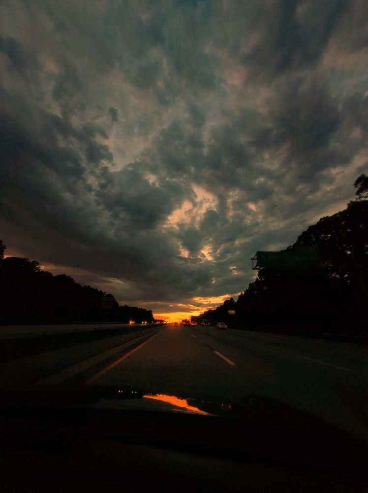 stormy sunset out the windshield of a car, road and treeline silhouetted against the breaking clouds & orange light
