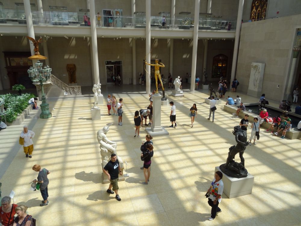 People walking through the Metropolitan Museum of Art American wing with the shadow of the glass atrium forming a grid on the floor. 