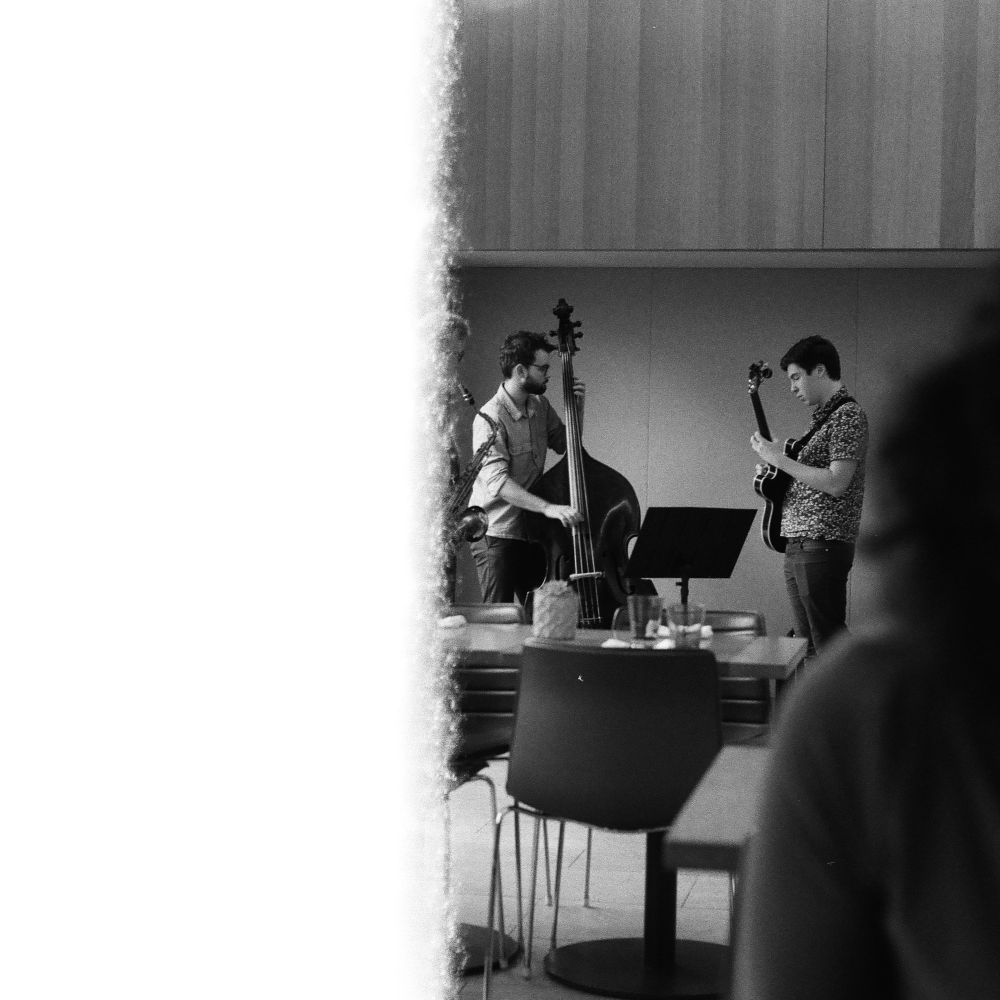 Half cut off end of film roll black and white photo of a three piece band playing for brunch audience within a museum space. The band is made up of a saxophone, guitar and a floor bass. 