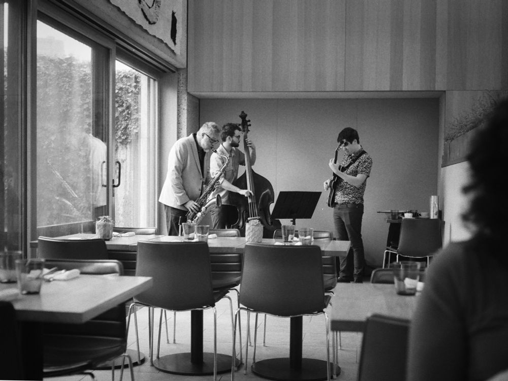Black and white photo of a three piece band playing for brunch audience within a museum space. The band is made up of a saxophone, guitar and a floor bass. 