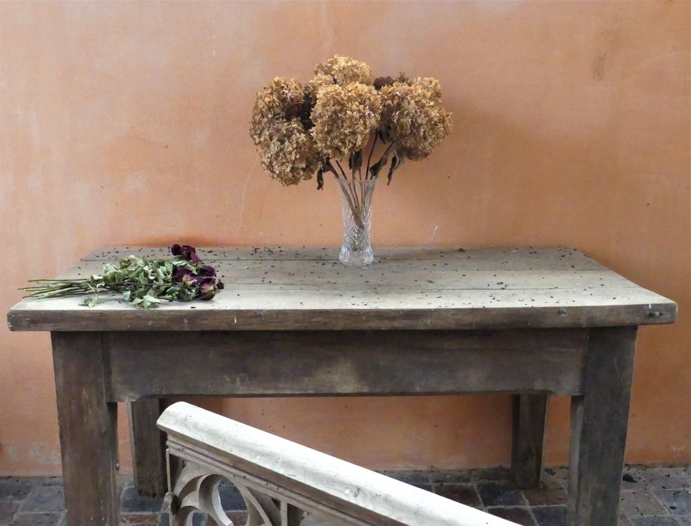 A vase with dead hydrangeas and a bunch of dead roses on a church table in front of a salmon-pink wall.
