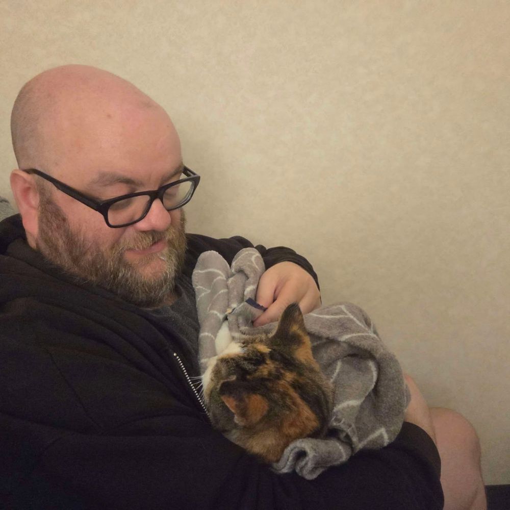 A picture of J.D. Lowe holding his cat, a young girl named Addie, from July 2025. She's swaddled in a blanket. We said goodbye to her that day.