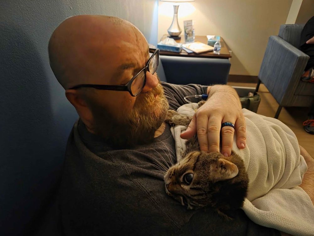 A picture of J.D. Lowe holding his cat, a young girl named Stalker, from February 2025. She's swaddled in a blanket; this is the night we said goodbye to her.