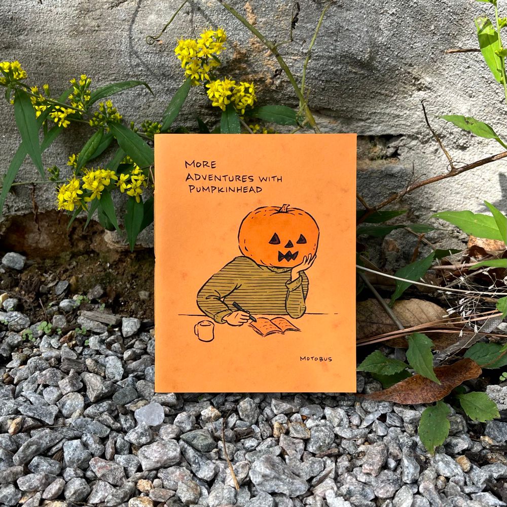 The photo shows the front cover to a mini comic, More Adventures with Pumpkinhead by motobus. Pictured on the cover: Pumpkinhead sits at a table. He is paused over a notebook — pen in hand. His chin rests on the other hand. He is thinking. A coffee cup sits at his side. The physical mini comic leans on an exterior wall with gravel below and wildflowers and weeds coming through the cracks.