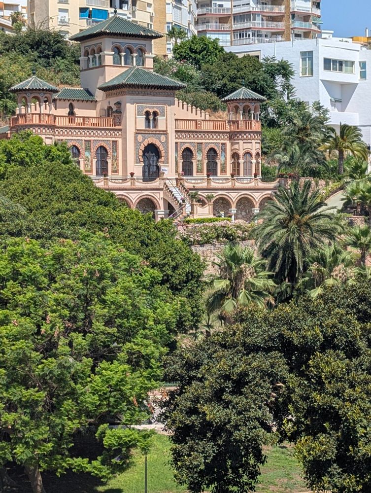 A photo of Casa de los Navajas in Torremolinos, Spain. It is a fairly opulent pink-brick building with multiple round arched windows and multiple low square pyramid roofs. It is built into a slope and surrounded by various lush Mediterranean foliage, including palm trees. It has a long stone staircase to its main door, possible cut in marble. It does indeed look like both a historic villa and a casino. It is apparently in the Moroccan style. 