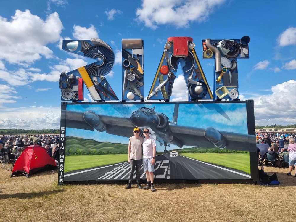 Photo infront of the RIAT logo at RAF Fairford for the Royal International Air Tattoo 2025. 