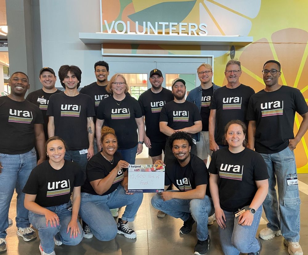 A group of URA employees wearing the organization t-shirt take a photo. Two of the employees hold a small sign that says "I helped sort 12,500 pounds of food."