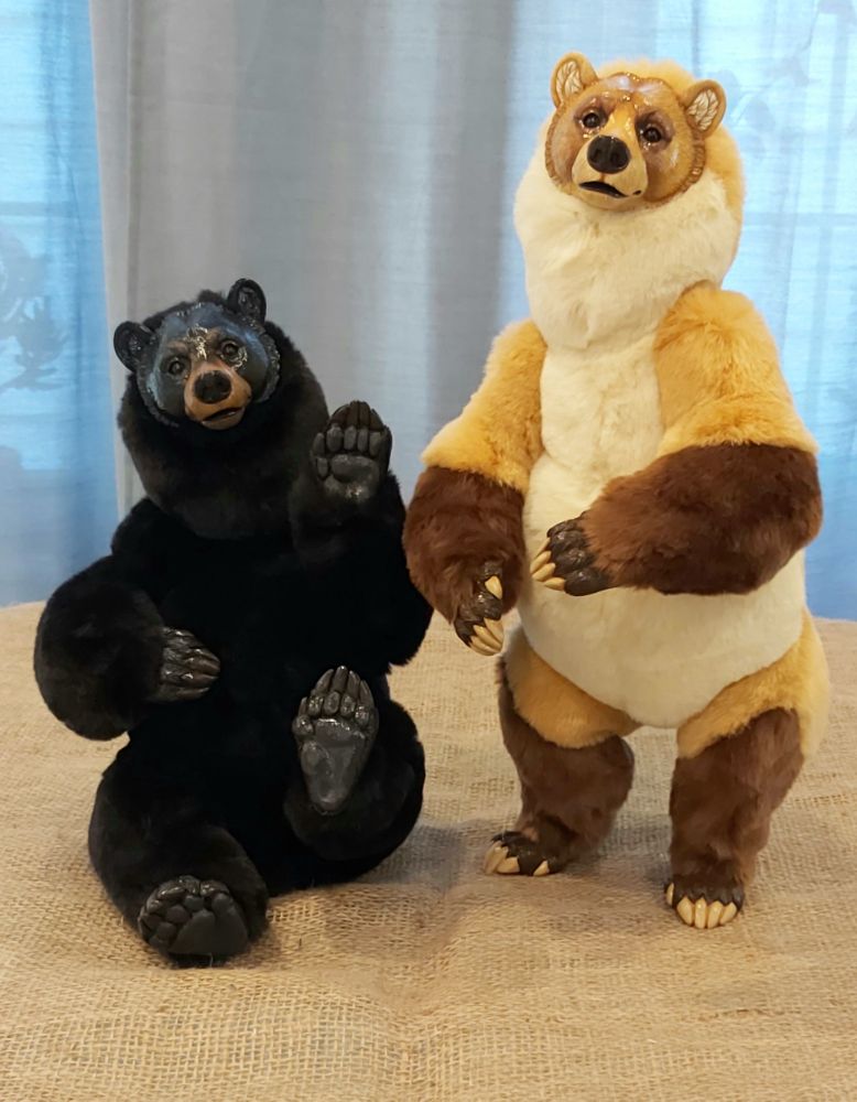 Two bear art dolls. One is a black bear, sitting with its forepaw uplifted, the other a golden grizz with darker socks standing on its hind legs. Both are made of rounded soft shapes with broad heads and dark eyes.