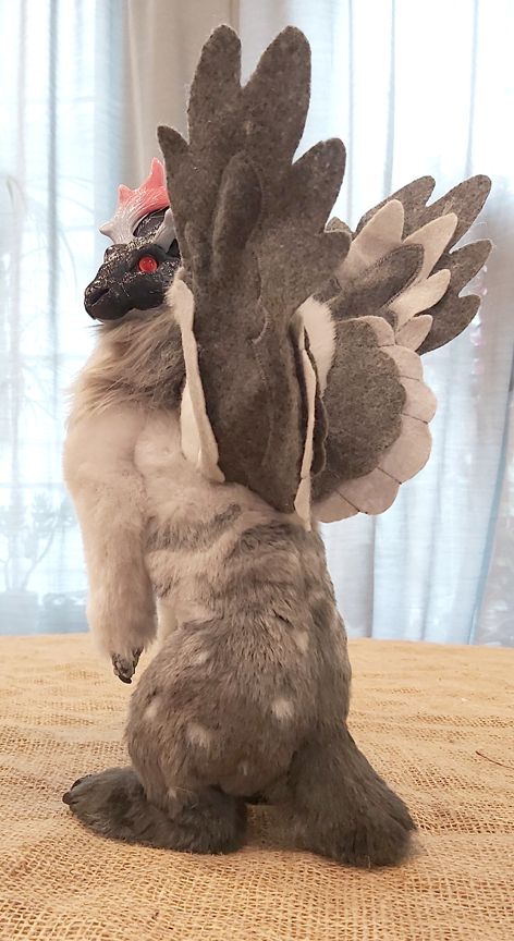 A winged hare art doll, with short branching antlers sits up facing away from the viewer, looking back over its shoulder. Its a series of soft greys with small pale grey spots on its haunches. Its antlers are a soft grey fading to a pink that matches its albino-pink eyes.