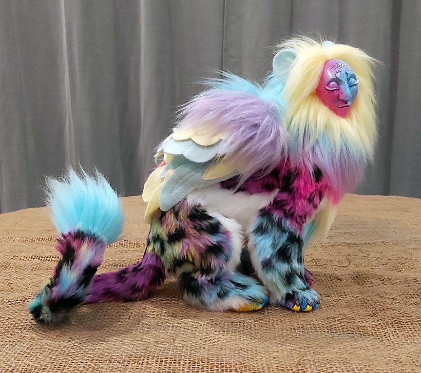 A multicoloured leopard sphinx art doll sits demurely, feet tucked together, wings folded. Her face is a blend of metallic magenta and blue, with closed eyes and a serene mask-like quality. Her mane is a muted rainbow, with blue lion ears sticking out, and her tail tip is curled.