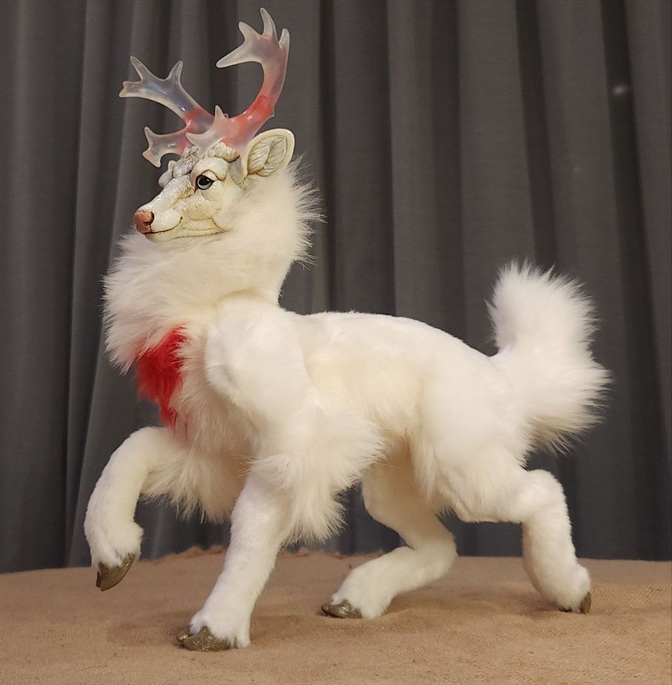 A white stag art doll, prances. Its body is a soft ivory with a thicker mane and a brilliant blood red patch at its throat. Its antlers are multi-tined, and translucent with a soft blue shimmer. Inside, are two swirled drops of heart's blood red. 