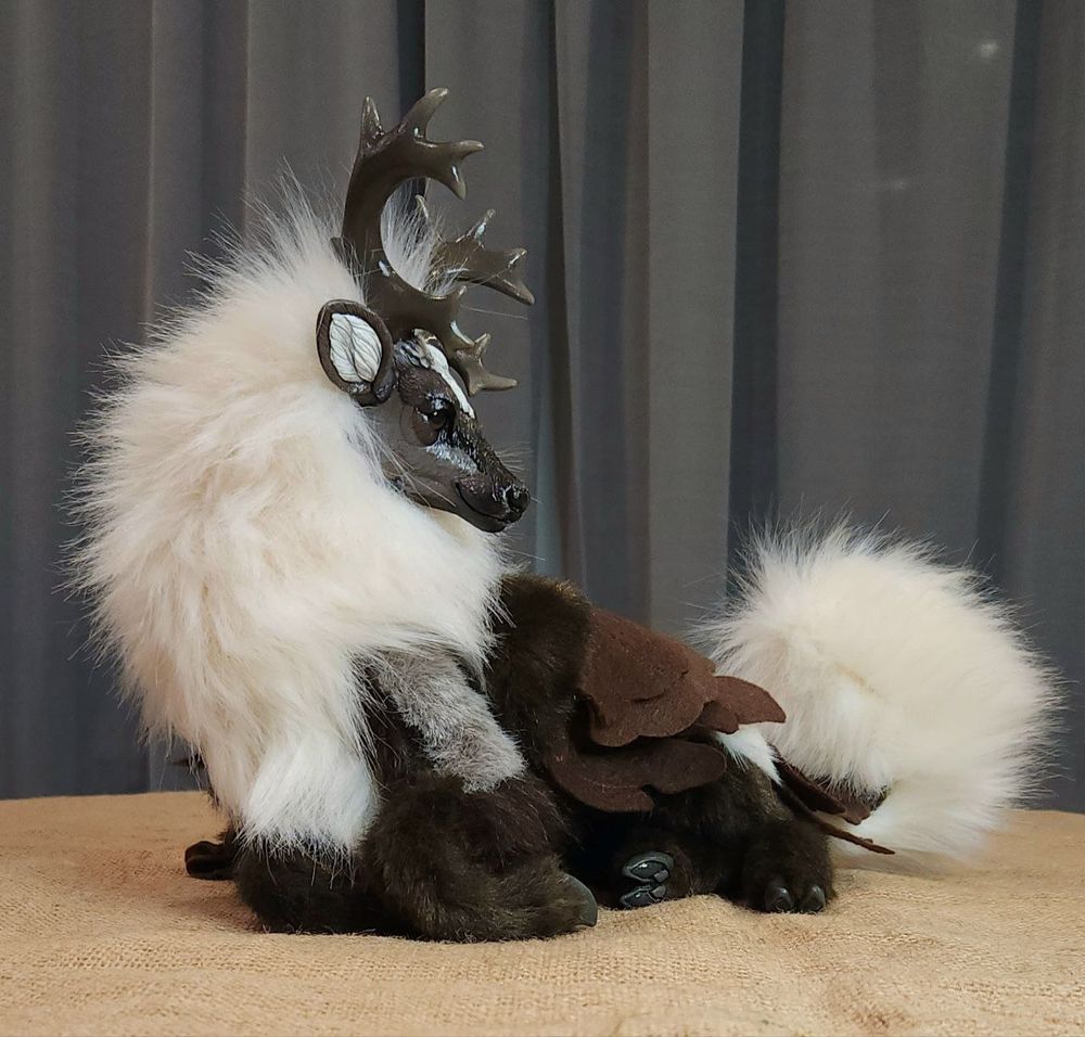 A peryton art doll laying down comfortable, front feet folded underneath them. They're caribou coloured with a thick ivory mane and dark walnut coloured legs, and a roan body. Their wings are folded flat to their sides, and their tail is curved gently.