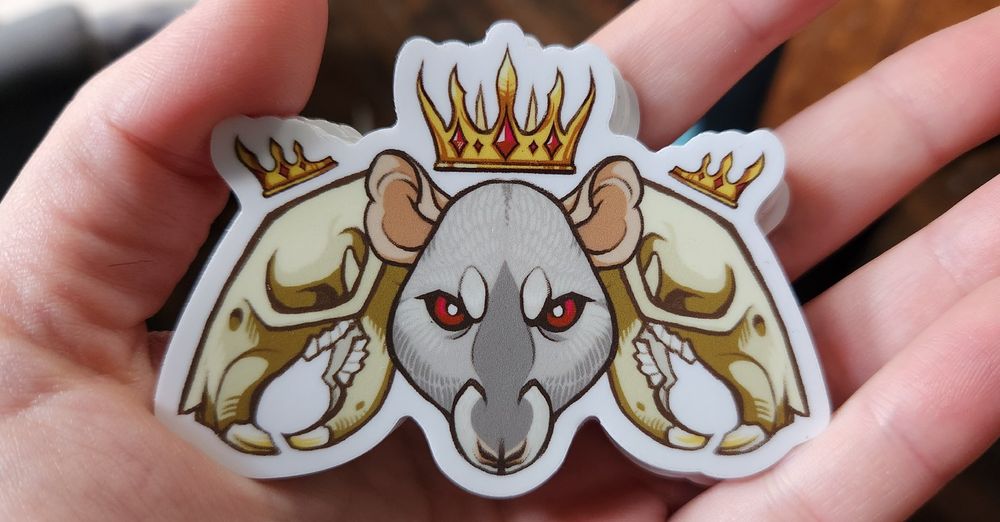 A sticker sits in my palm. In the center is a grey rat face, red eyed and crowned. To either side is a rat skull with its own smaller crown.