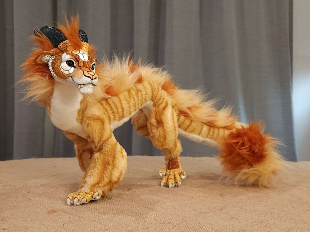 An orange tabby dragon art doll stands looking left. Its face has large cream blocks of scales, with a soft orange gradient from muzzle to ears that matches its striped fur. Its horns are a dark greeny-grey, translucent with just a hint of sparkle.