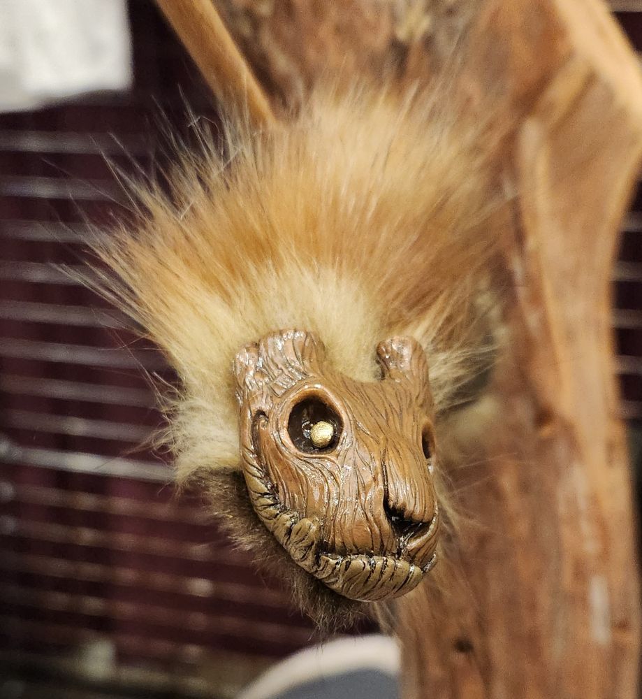 The face of a small art doll with wood texture, amd gold eyes that looks like a smiling stump.