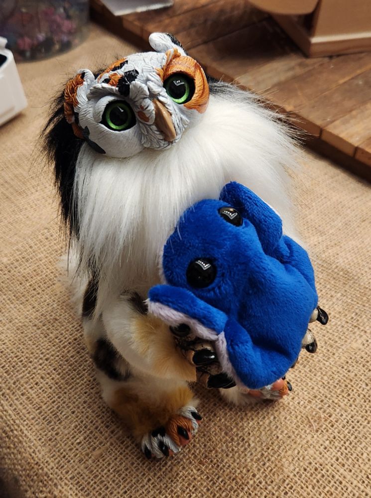 A small calico art doll of an owl gryphon kitten holding a small blue plush frog.