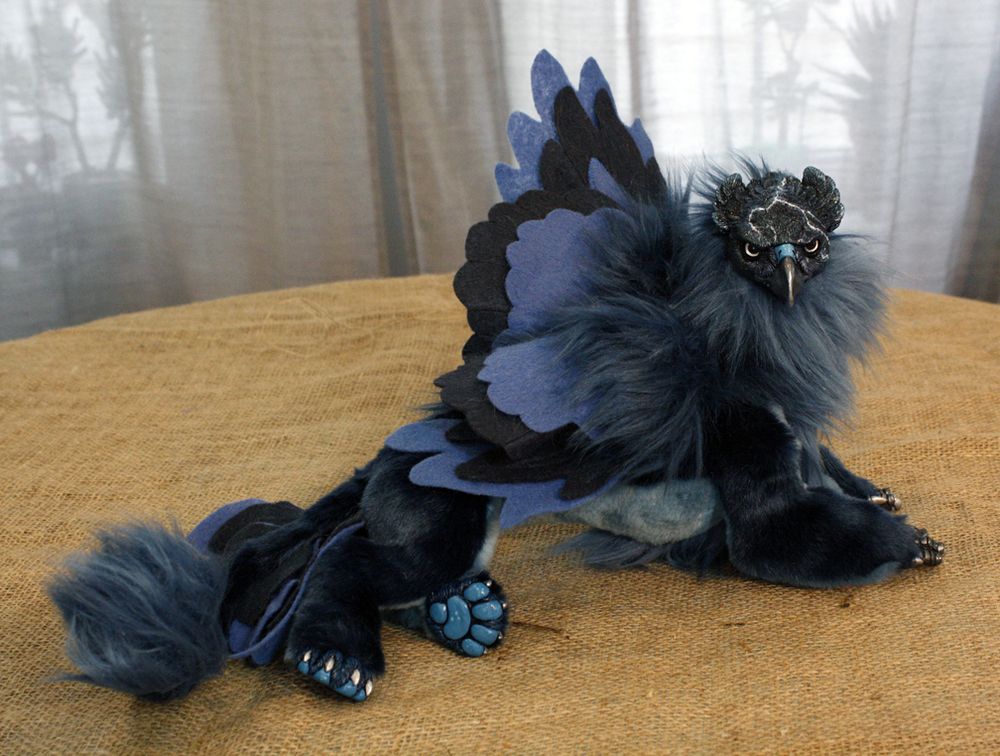 A dark, stormy coloured gryphon art doll lies with wings partially lifted. Their head is turned to face the viewer, showing a face streaked by lightning-like markings and bright silver eyes. Their body is made of soft furs in moody grey-blues.