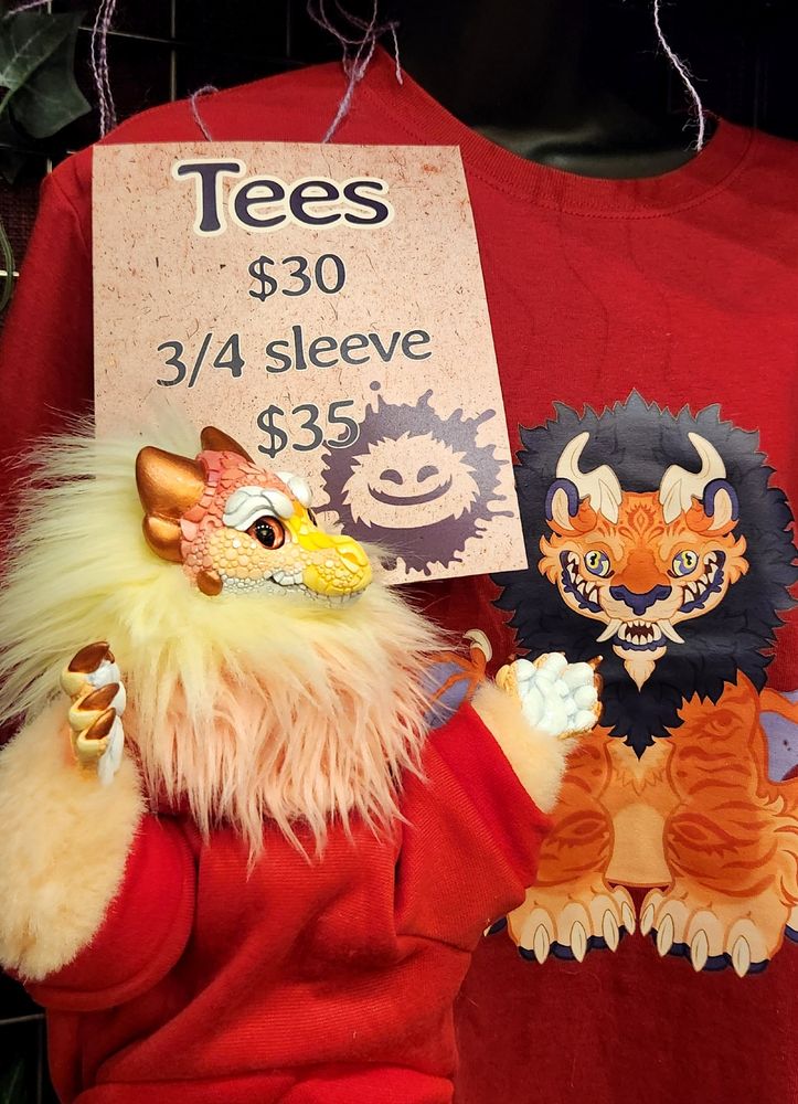 A small dragon art doll in a red hoodie stands beside a red shirt with a manticore on it.