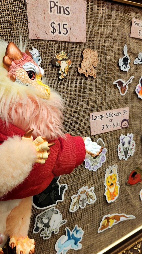 Peaches the dragon (art doll) stands showing off a board of stickers

