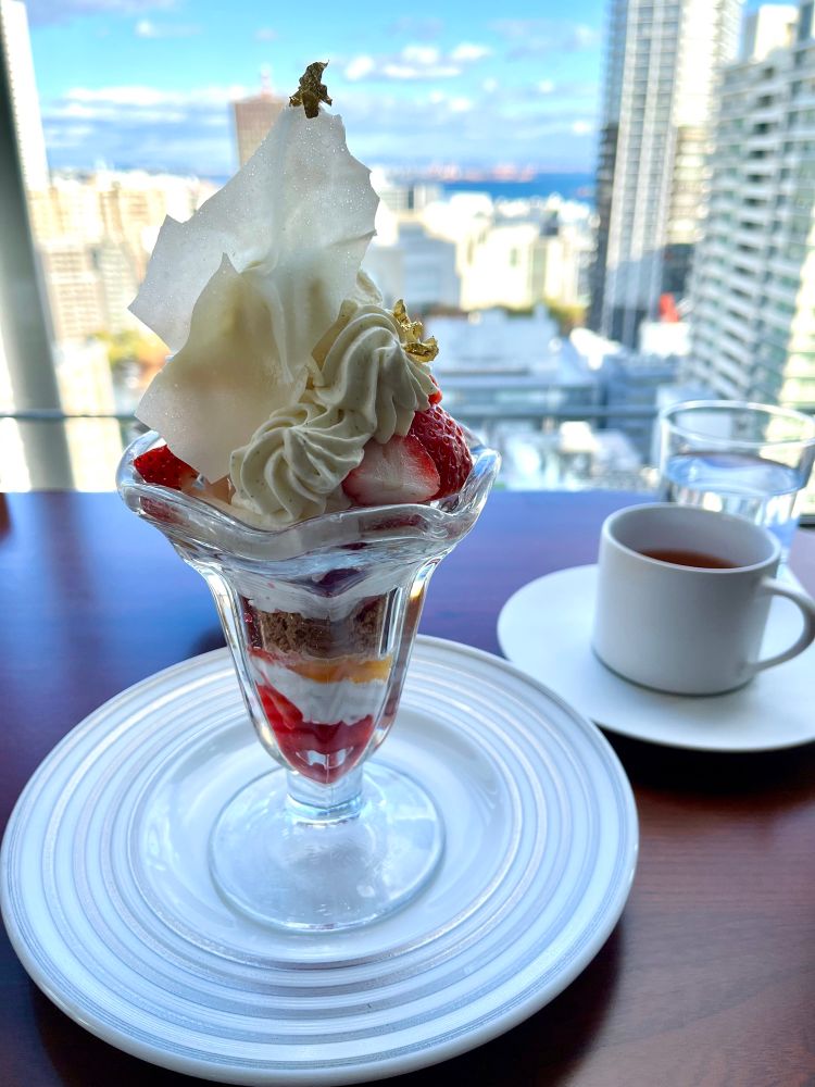 手前に苺のパフェ、右手に紅茶のカップとソーサー、パフェ越しに街の眺望が映った写真。