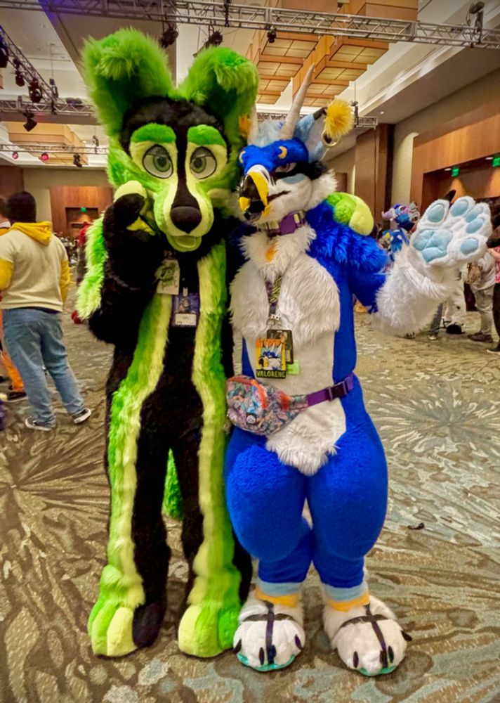 A fursuiter named Lintrap (black and green canine) stands next to me as Val. (blue avian) We are each have an arm wrapped around eachothers backs. Lintrap has a paw to their snout in a cute pose, and I have an open paw stretched out to the side.