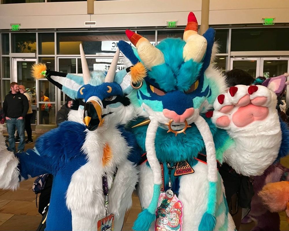 I'm fursuiting as Val (blue avian) while posing together with a blue dragon × feline hybrid fursuiter. We're shoulder-to-shoulder and both have a paw raised to pose.