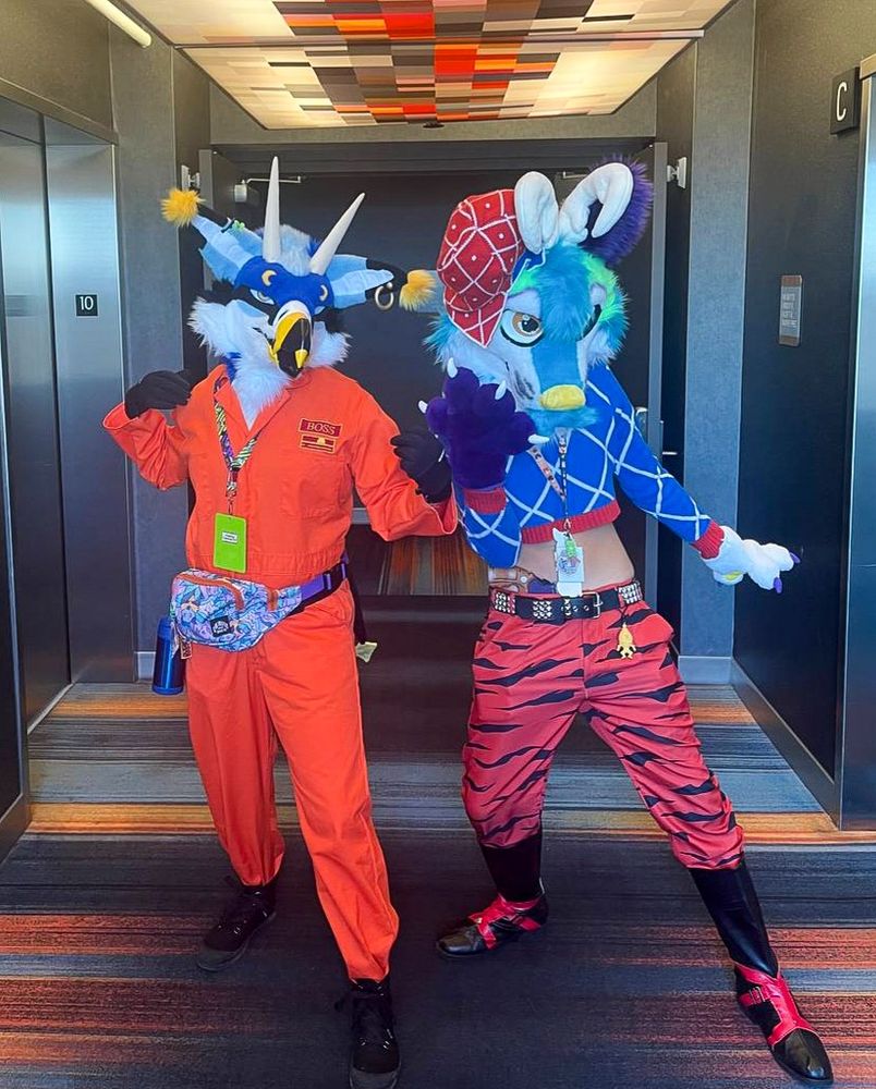 I'm geared up in my Lethal Company costume with Val's (blue avian) fursuit head on. Next to me is a friend cosplaying Mista from JJBA and wearing my sona, Ripple's, head. Ripple is a purple, blue, and green antlered wolf. My friend is striking Mista's signature pose, and I mimiced him.
