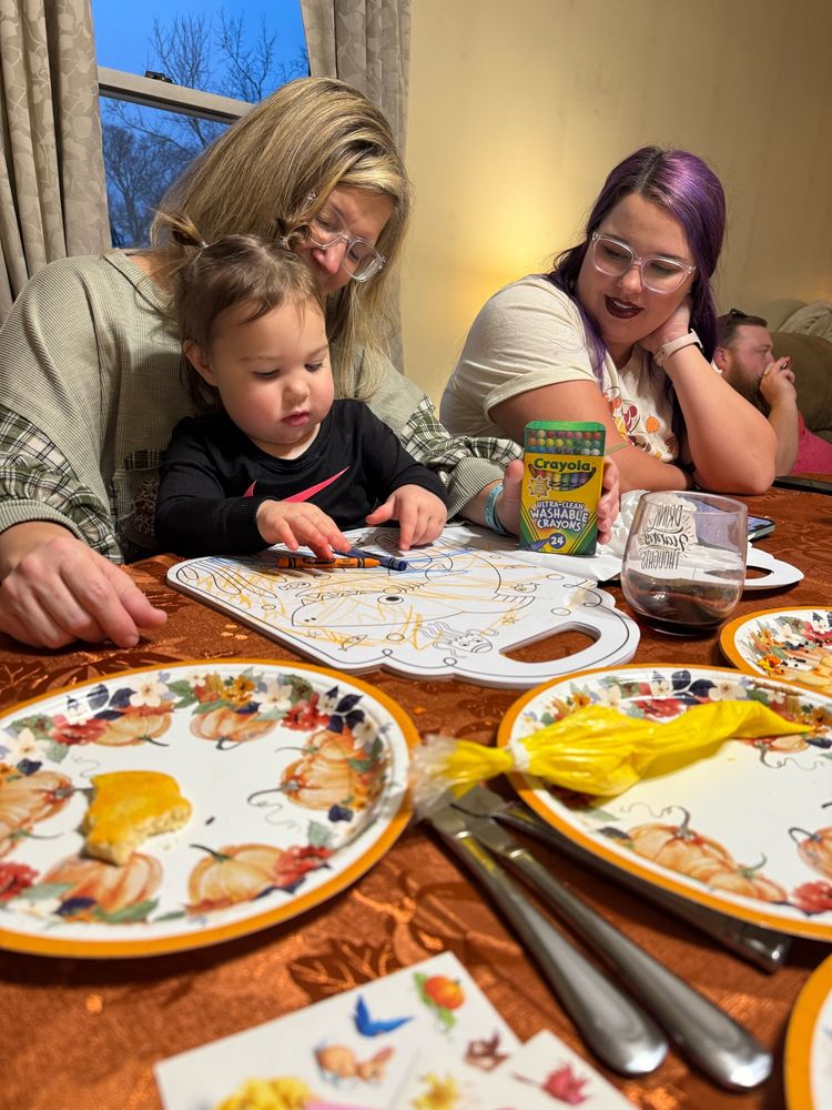 Toddler sitting on her grandma’s lap coloring during next to her mom looking over. 