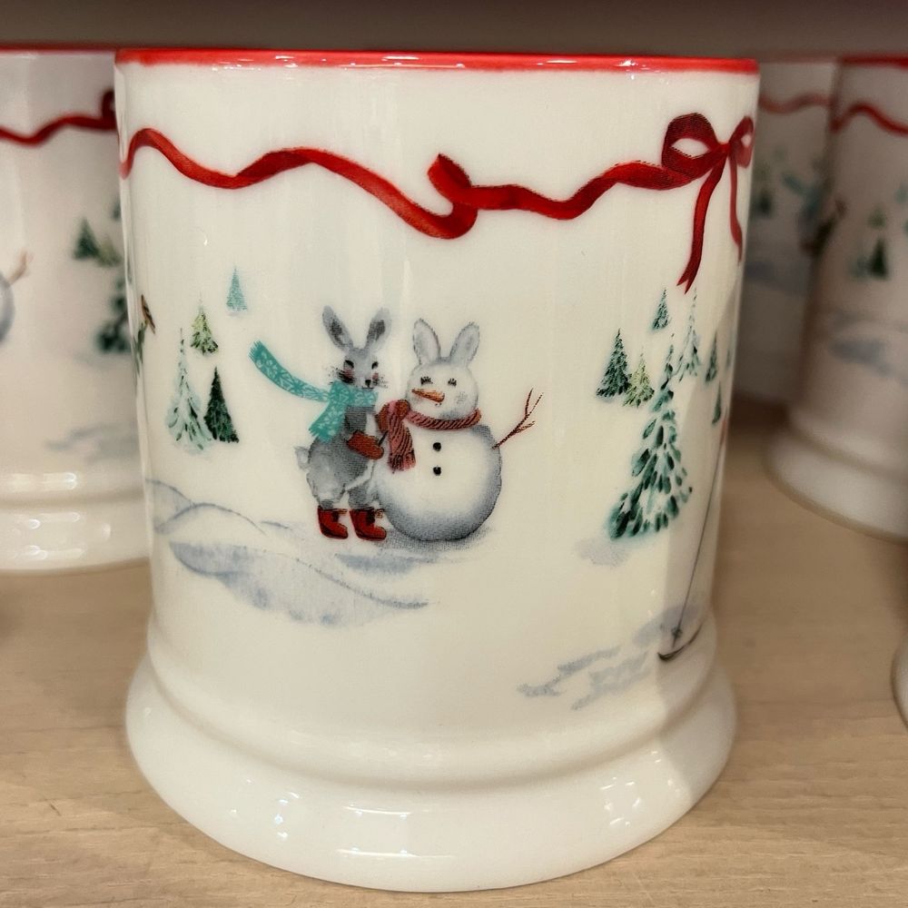 A festive mug with a hare in a green scarf making a snow bunny. There is a snow background with conifers with snow.