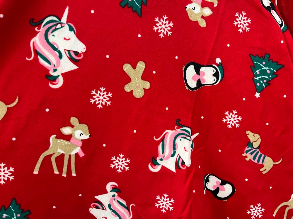 Festive red fabric decorated with stylized images of Yule trees, deer in scarves, gingerbread men, snowflakes, unilcorns (!), and penguins.