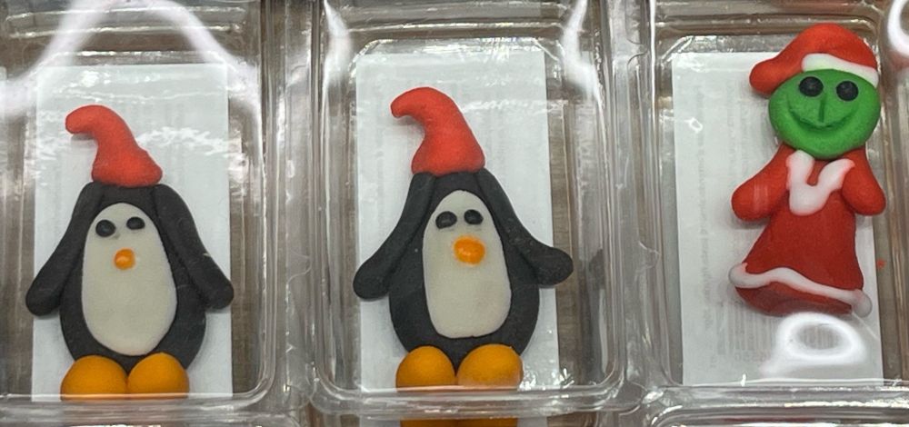 Two marzipan penguins wearing Santa hats next to a marzipan grinch. Each is stored in a small clear plastic container.