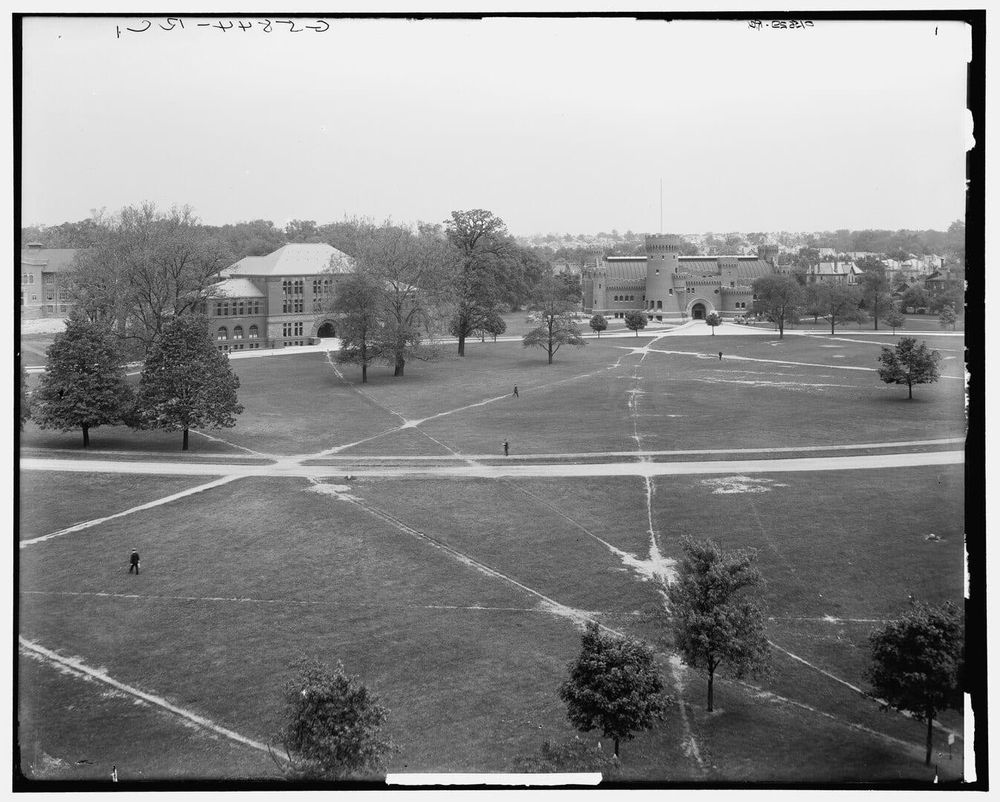 A photo from high ground showing a green between buildings at a university, with some paved paths and a myriad of unpaved clearly visible, downtrodden, highly used paths.