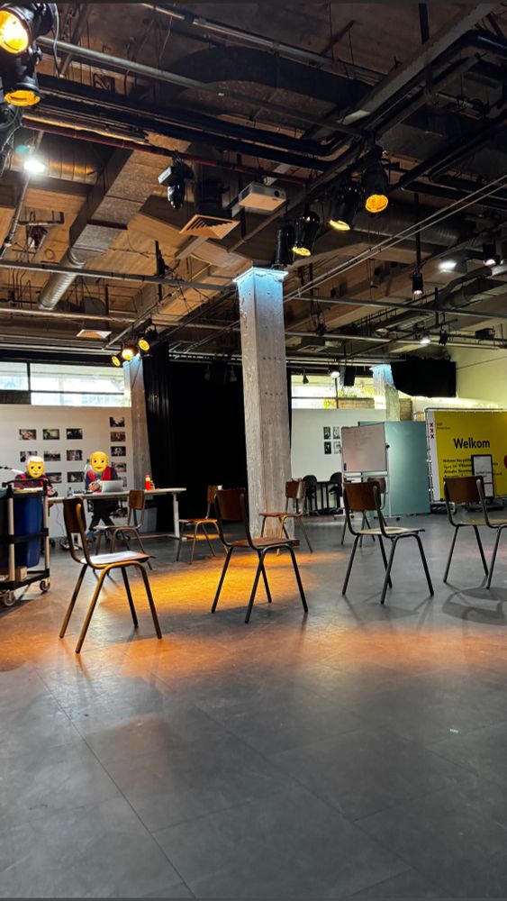 Auf dem Bild ist ein Raum mit industriellem Stil zu sehen – die Decke ist offen, sodass Rohre, Kabel und Beleuchtung sichtbar sind. Der Raum scheint für Veranstaltungen, Workshops oder Präsentationen oder Clubveranstaltungen genutzt zu werden.

In der Mitte stehen mehrere einfache Holzstühle mit Metallbeinen, die in lockeren Abständen zueinander aufgestellt sind. Der Boden besteht aus dunklem Material, wahrscheinlich Linoleum oder glattem Beton.

Im Hintergrund gibt es Tische mit Computern, auf denen zwei Personen mit gelben Emoji-Gesichtern sitzen. An der Rückwand hängen mehrere Fotos oder kleine Poster. Rechts im Bild befindet sich eine große gelbe Wand oder ein Aufsteller mit dem Wort „Welkom“, was darauf hindeutet, dass der Ort in den Niederlanden liegt.

Die Beleuchtung im Raum ist warm und teils auf bestimmte Bereiche gerichtet – offenbar handelt es sich um Bühnen- oder Studiolicht. Insgesamt wirkt der Raum modern, funktional und kreativ, wahrscheinlich Teil eines Bildungs- oder Kulturzentrums.