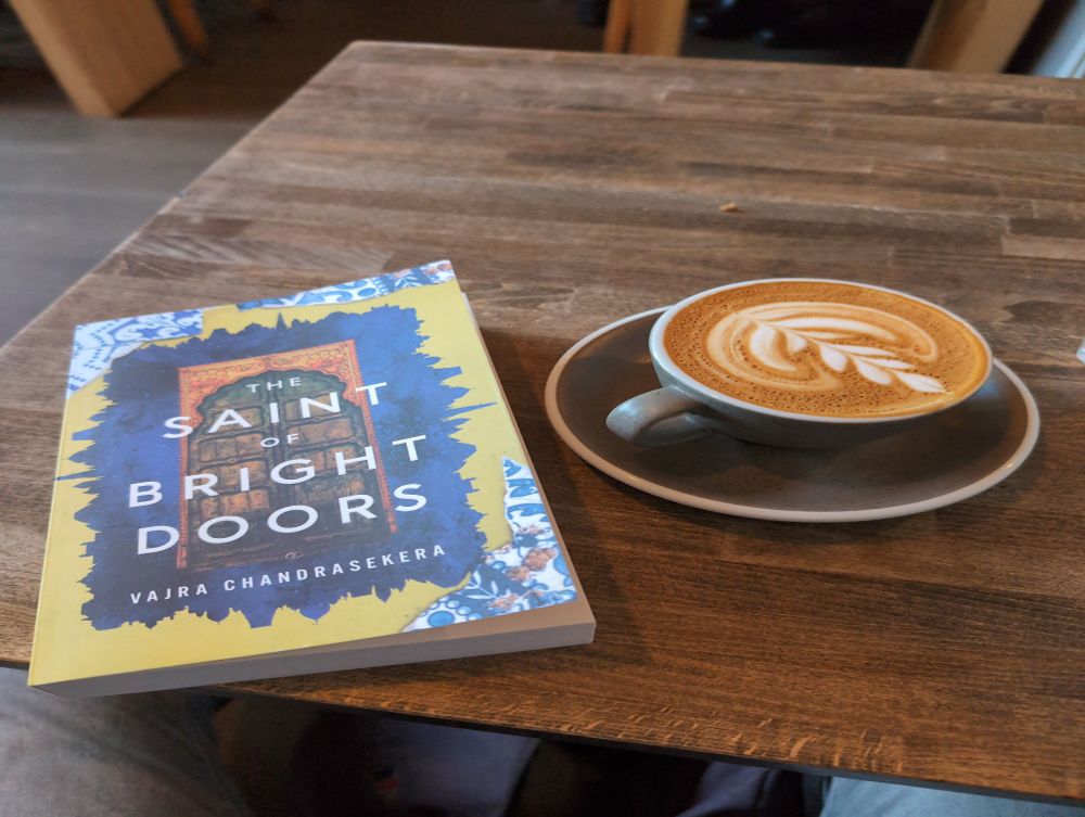 Novel "The Saint of Bright Doors" on a table next to a flat white