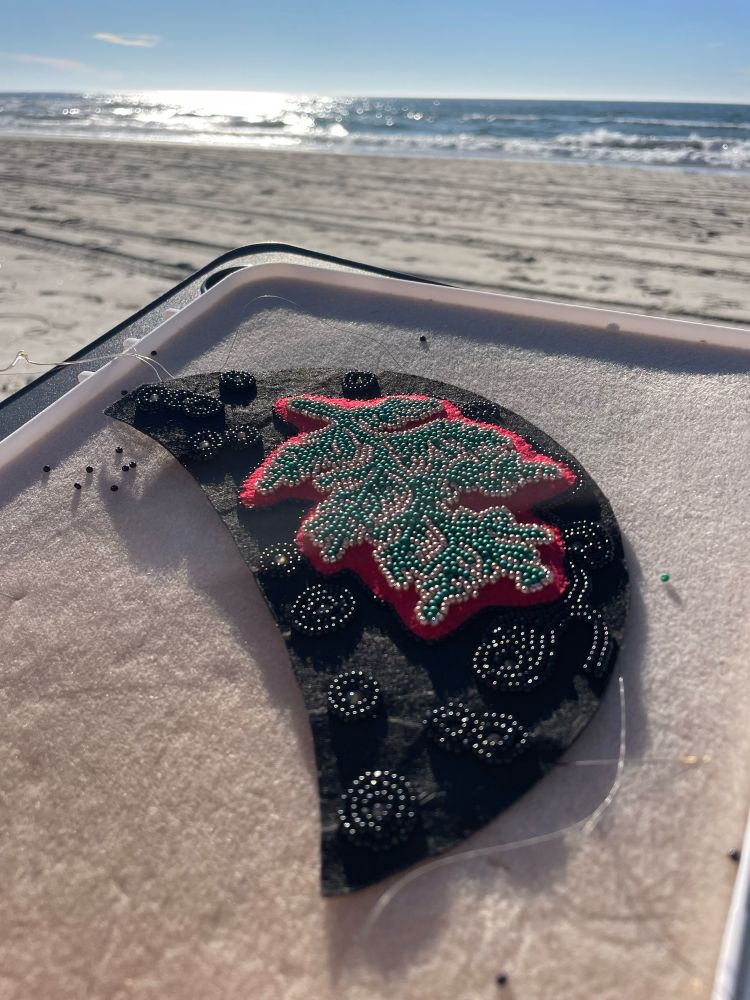 Black half moon shaped beadwork with a beaded cedar branch sitting on a beading tray with the ocean in the background. 