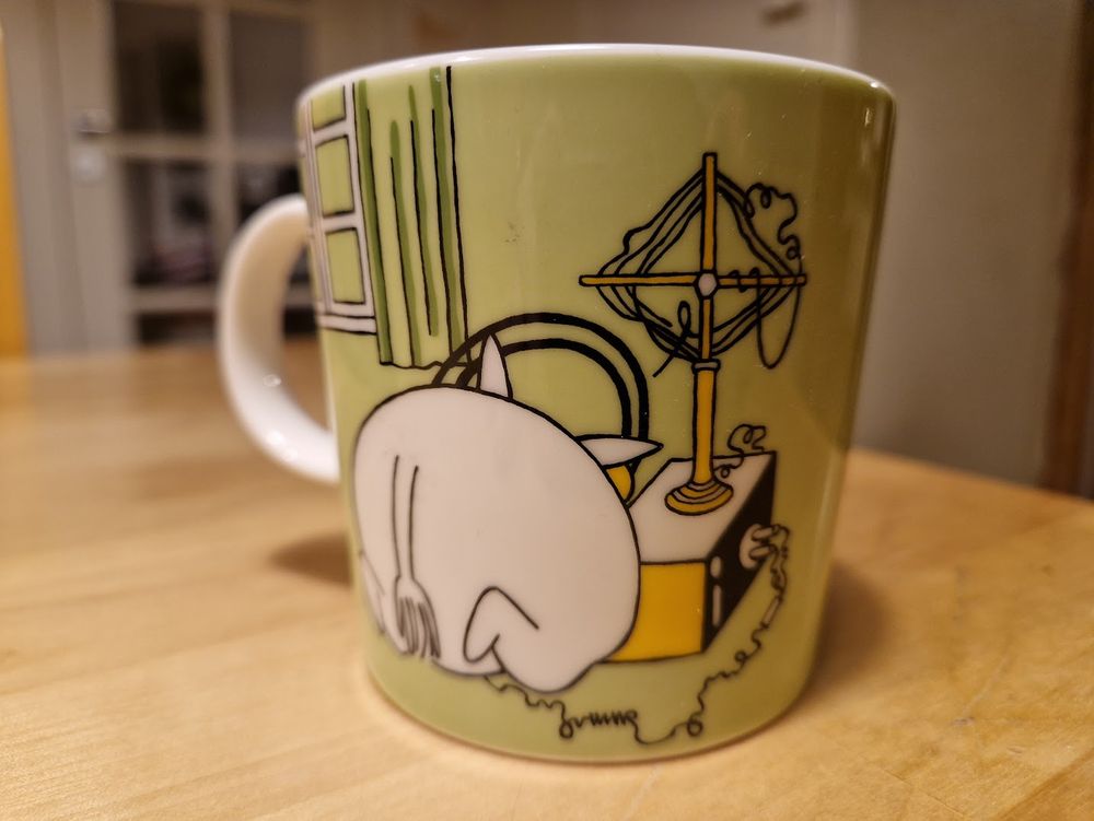 Vaalenvihreä muumikuppi, missä Muumipeikosta näkyy takapuoli, kun hän on kumartunut radion eteen. Radion päällä mielenkiintoisen näköinen antennihässäkkä.