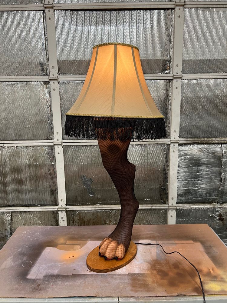A cursed lamp. The lamp is shaped like a hyena leg. The leg is wearing a thigh-high fishnet legging. The lampshade atop the leg has black fringe. This view shows the spots on the outside of the thigh.
