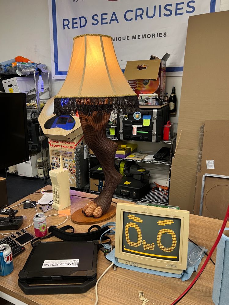 A scene within a hackerspace. The scene depicts a table with a laptop, a serial terminal, and a lamp. The laptop is ruggedized. The terminal has an amber display showing an angry "owo" face. The lamp is shaped like a hyena leg. The lamp is wearing a thigh-high fishnet.