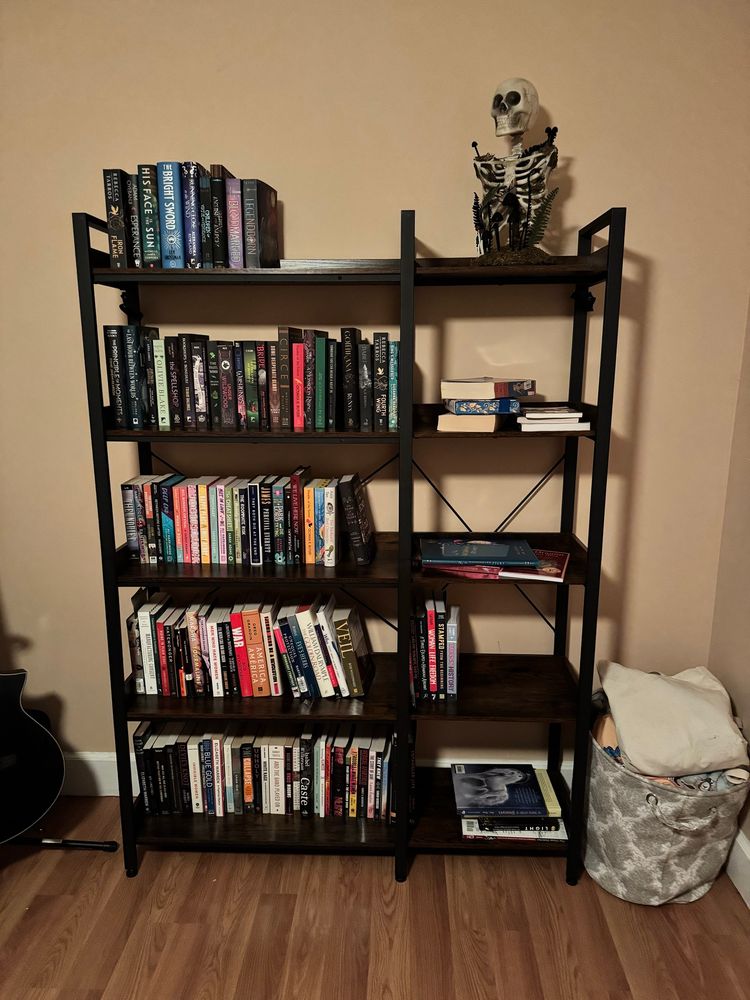A wood and metal bookshelf with five shelves, which are partially filled with books. On the top right, there’s a faux skeleton with just a skull and rib cage with moss and mushrooms on it, to appeal to my latent bog witch tendencies. The wall behind the book shelf is, sadly, beige. 