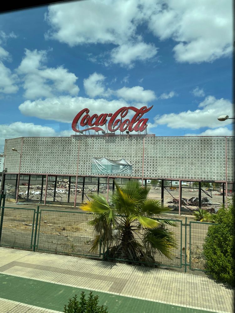 Zerfallenes Gebäude mit Coca Cola Schriftzug, davor eine kleine Palme, darüber blauer Himmel mit Wolken.