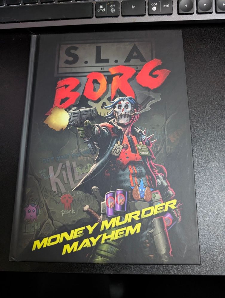 A photo of the SLA BORG TTRPG book on a black desk. The cover features a character wearing a skull mask and shooting a gun, with a damaged wall, covered in graffiti behind them.