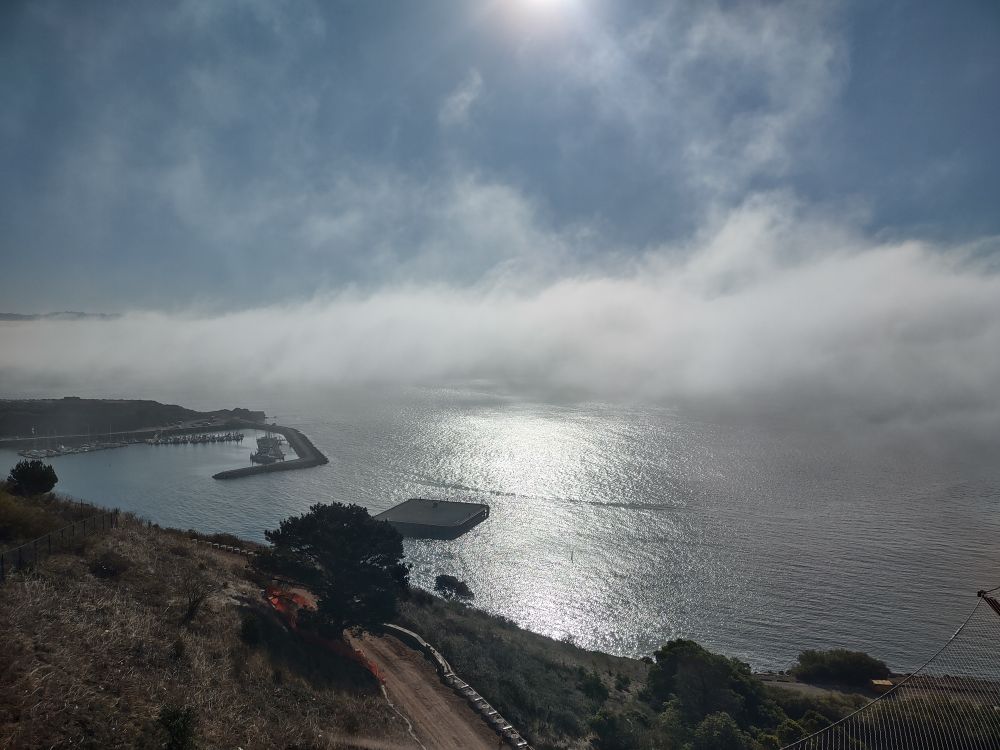 Lower part of the image is a bit of dirt road and dry brush with a couple of trees giving way to a silvery bay with a bright stripe of sun reflection; a thick marine layer is against the horizon with huge tufts wafting up into the blue sky; a bit of land and a thin dock jut out from the left side of the frame