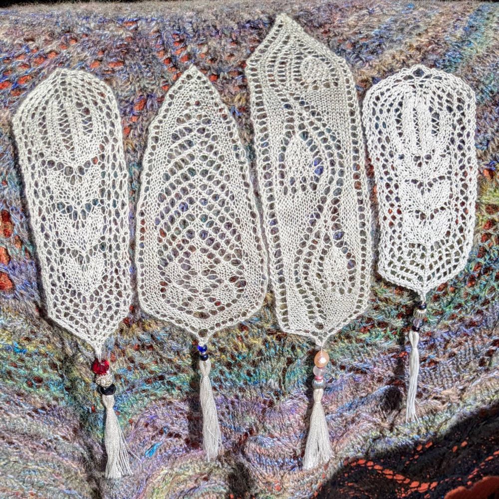 Four white lace bookmarks, each finished with beads and a tassel. One gas a tulip motif and red silver and black beads. The next is like a large leaf, with blue beads, and the next is like snow drop flowers with pink beads. The last is another tulip, though smaller, with purple beads.

They are on a background of a knitted lace shawl in shades of purple and green.