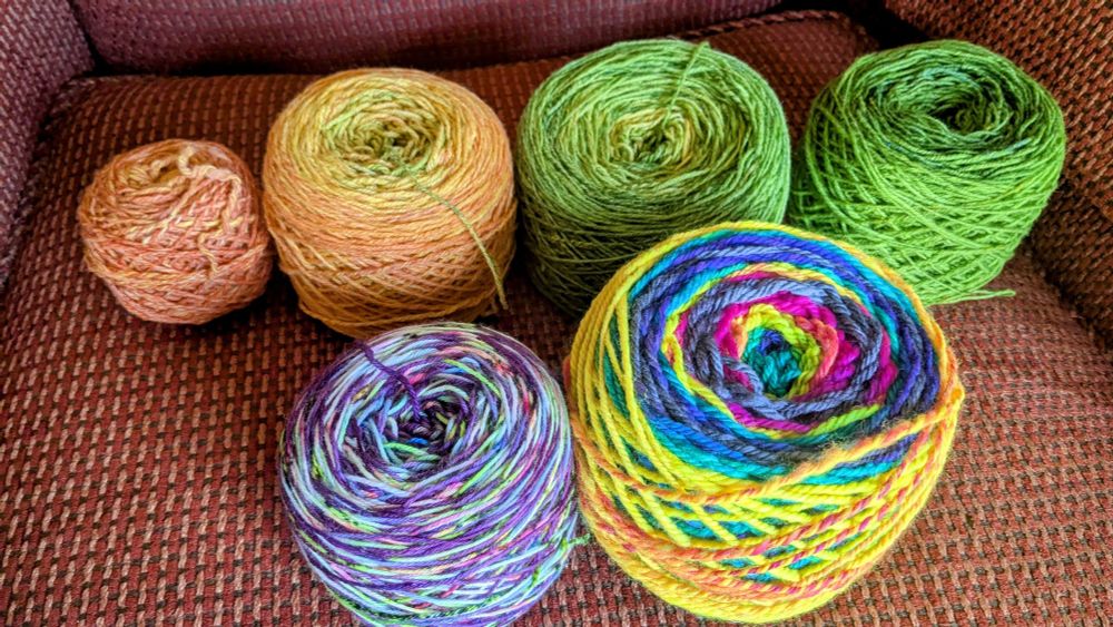 Six cakes of yarn sitting on a chair. Top row is a gradient from one orange cake, to a yellow orange, to a yellow green, to a green. Bottom left is sock yarn in purple with spots of highlighter yellow orange and pink. Bottom right is chunky yarn in neon rainbow colors with grey in between each repeat.