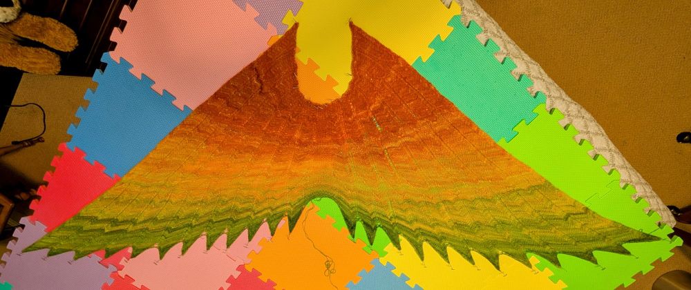A knitted shawl shaped like outstretched wings pinned to a foam mat on a bed. The shawl is a gradient from orange to yellow to green at the feather tips. It's probably five feet wide!