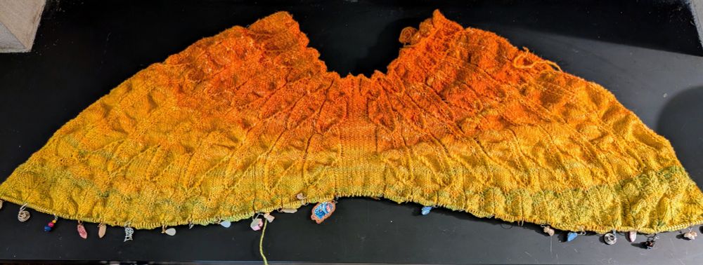 A knitted shawl in progress. A gradient from orange at the top to yellow to yellow-green at the bottom. (The remaining yarn, not pictured, transitions to green) The top edge of the shawl is a semicircle, and it spreads out into two half formed wings. The stitches are arranged to look like feathers, but it's still wrinkled and scrunched in texture. The bottom edge is still on a long knitting needle, with a couple dozen stitch markers separating every feather repeat. Some of the markers are shaped like birds, and there is a silver spinning wheel charm and a blue sheep as well.