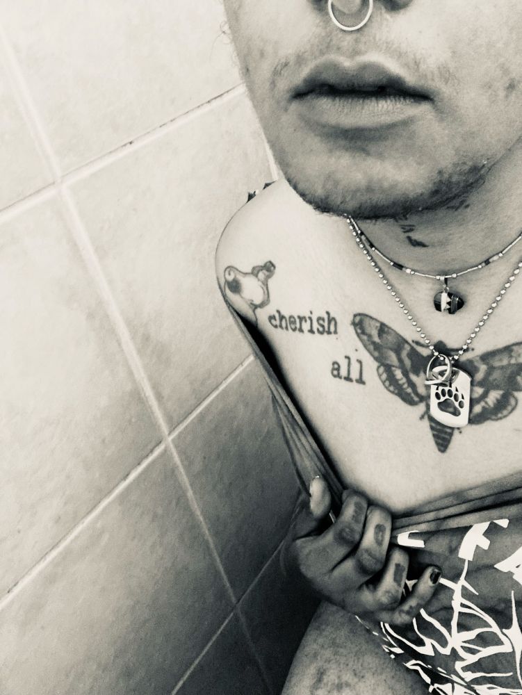 black and white selfie of cain in a low cut t-shirt that they’re pulling down to show more skin. they’re heavily tattooed and wearing silver jewelry in their nose and around their neck. their mouth is slightly open and they have the whispy beginnings of a moustache and beard.